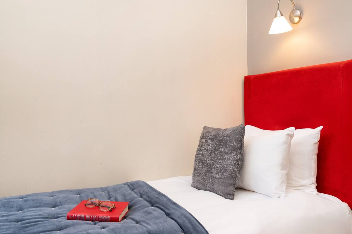 A neatly made bed with a red headboard, a grey and white pillow, and a red book with glasses on top.
