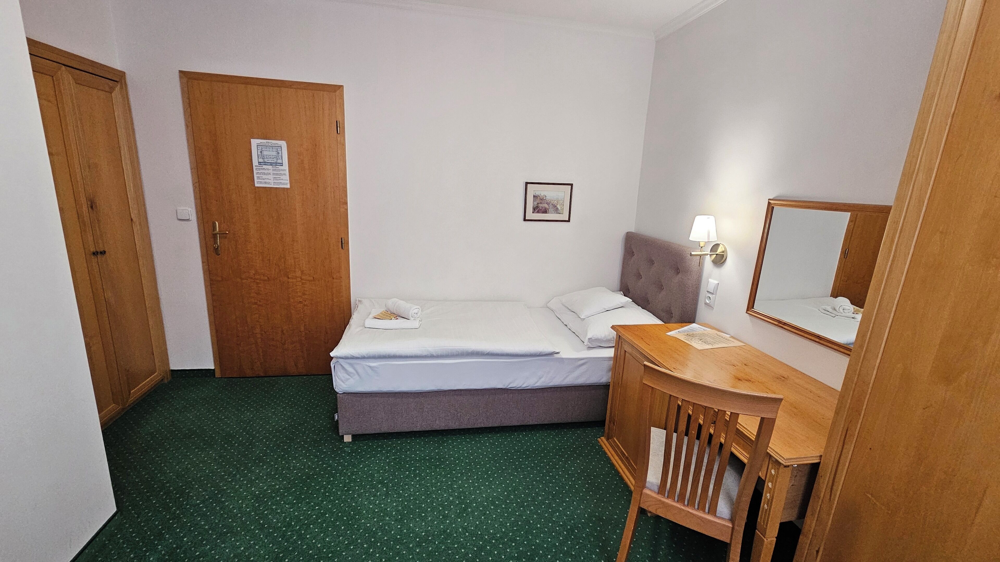 Quarto de hotel com cama, escrivaninha de madeira, espelho, luminária na parede e porta.