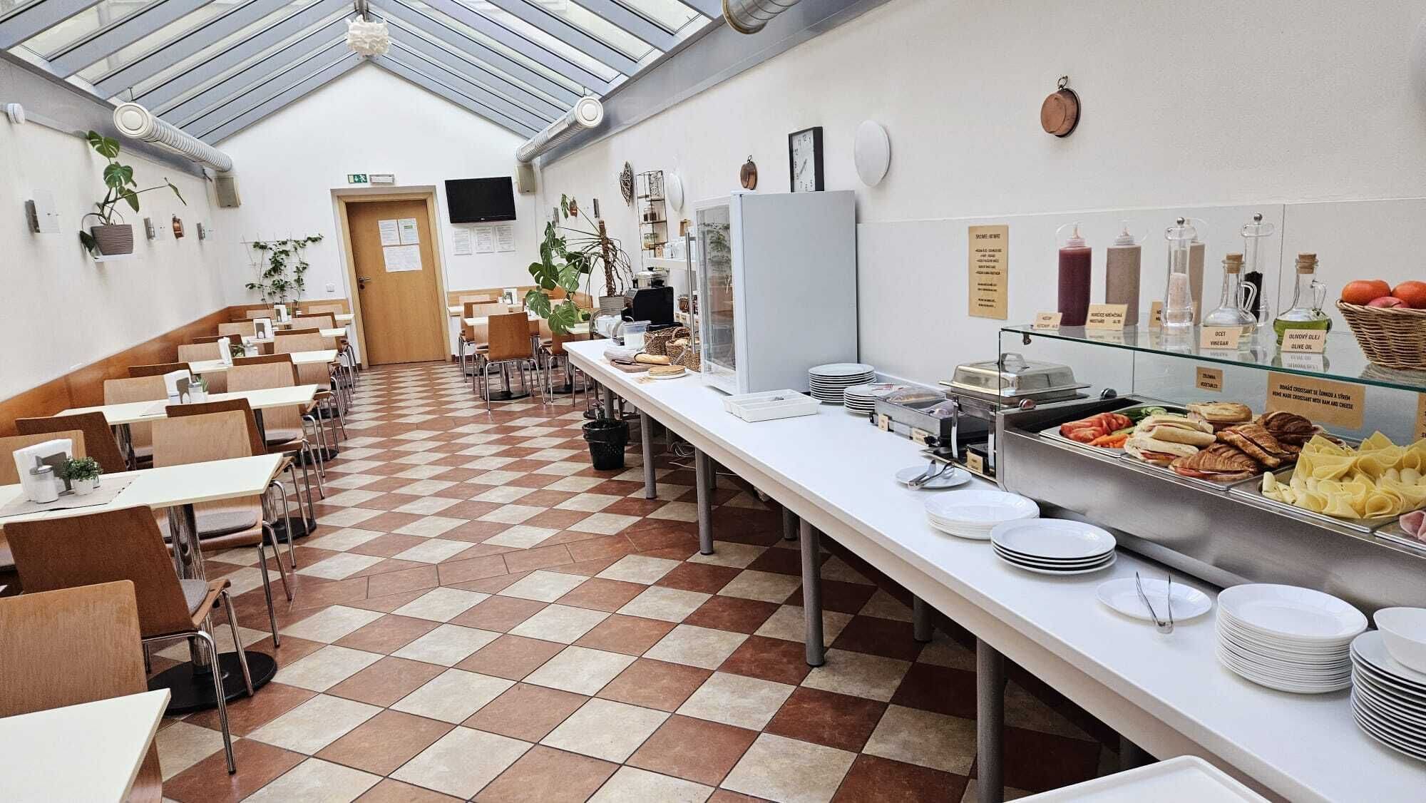 Una cafetería con piso de baldosas de cuadros, varias mesas y sillas, un mostrador de vidrio con alimentos y un techo alto con tragaluces.