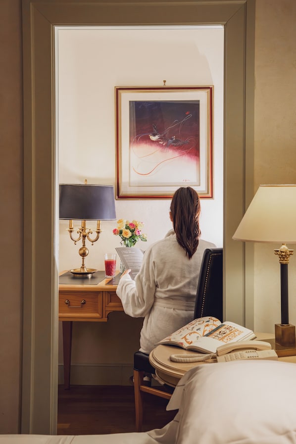 A hotel room with a desk, a chair, a bed, a lamp, a framed picture on the wall, and a vase with flowers.