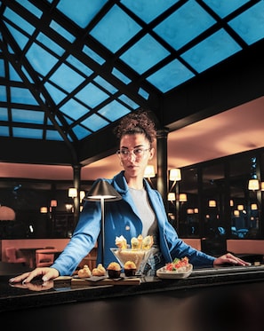 A person standing at a counter with a glass display of desserts, in a room with a high ceiling and large windows.