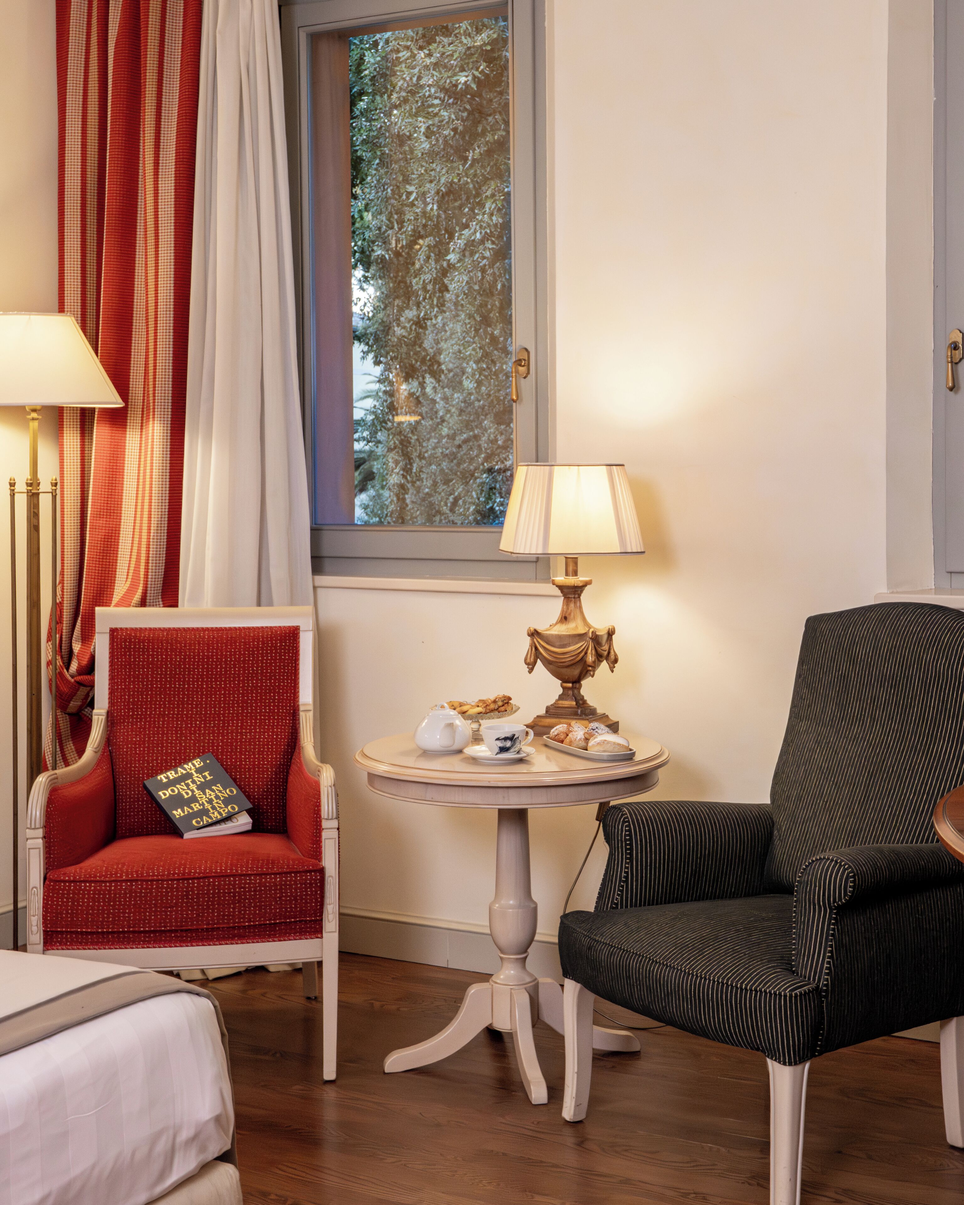 A room with a red armchair, a black armchair, a small round table with a lamp, and a window with a view of trees.