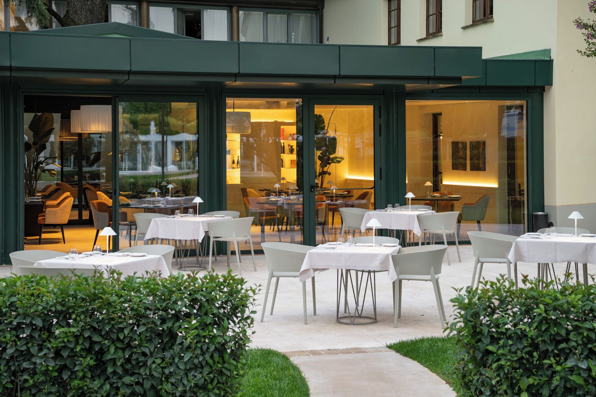 An outdoor dining area with tables and chairs, surrounded by greenery and a building with large windows.