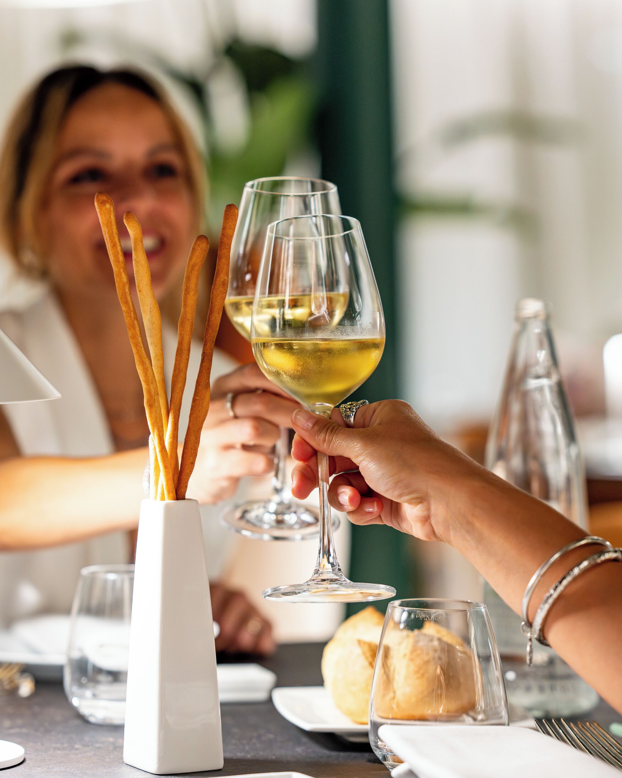 A dining table with a white vase containing breadsticks, a glass of white wine, and a bottle of water.