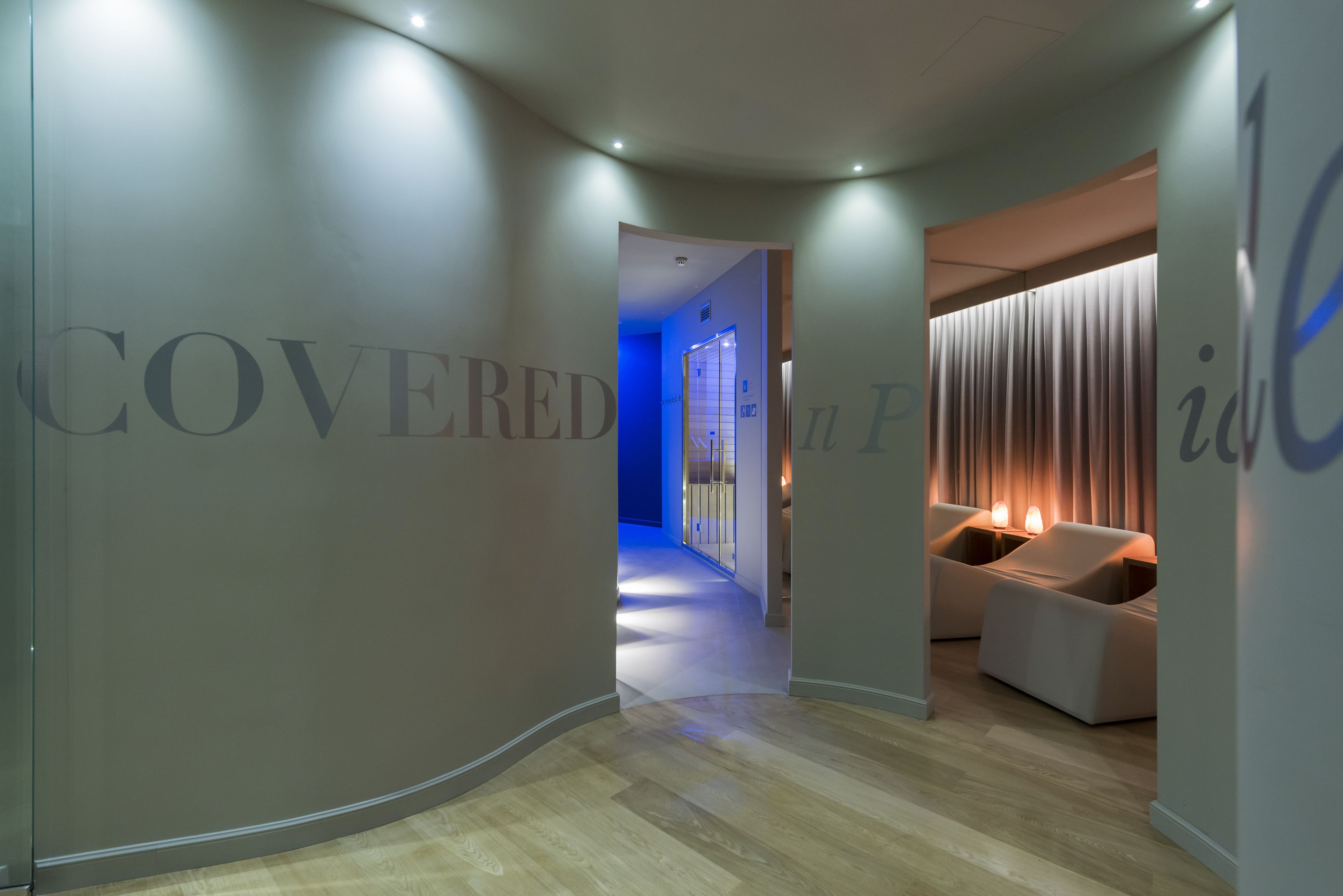 A modern hotel corridor with a wooden floor, a blue-lit hallway, and a seating area with a lamp.