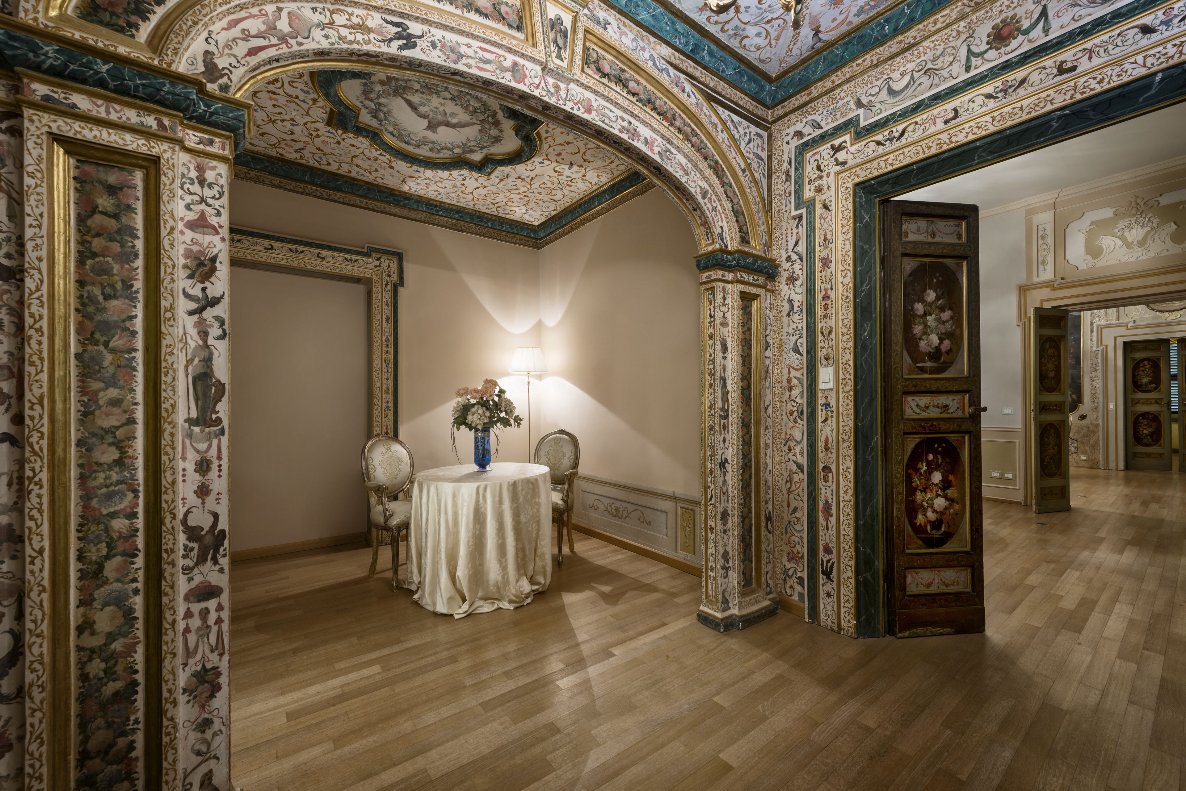 A room with ornate ceiling and wall decorations, a round table with a vase, and a lamp.