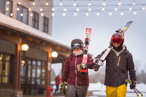 Two people holding skis, walking in a snowy area with buildings in the background. - Hôtel Stoneham (Stoneham-et-Tewkesbury)