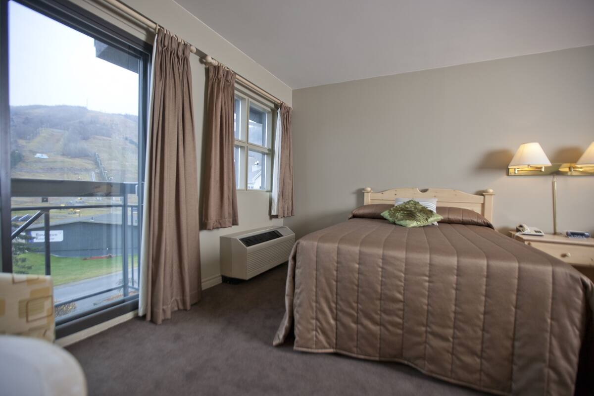 A bedroom with a large window, an air conditioning unit, a bed with a brown bedspread, a nightstand with a lamp, and a view of a building and landscape outside.