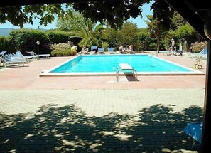 Una piscina rectangular con un pequeño trampolín en el centro, rodeada de sillones de descanso y una terraza pavimentada.