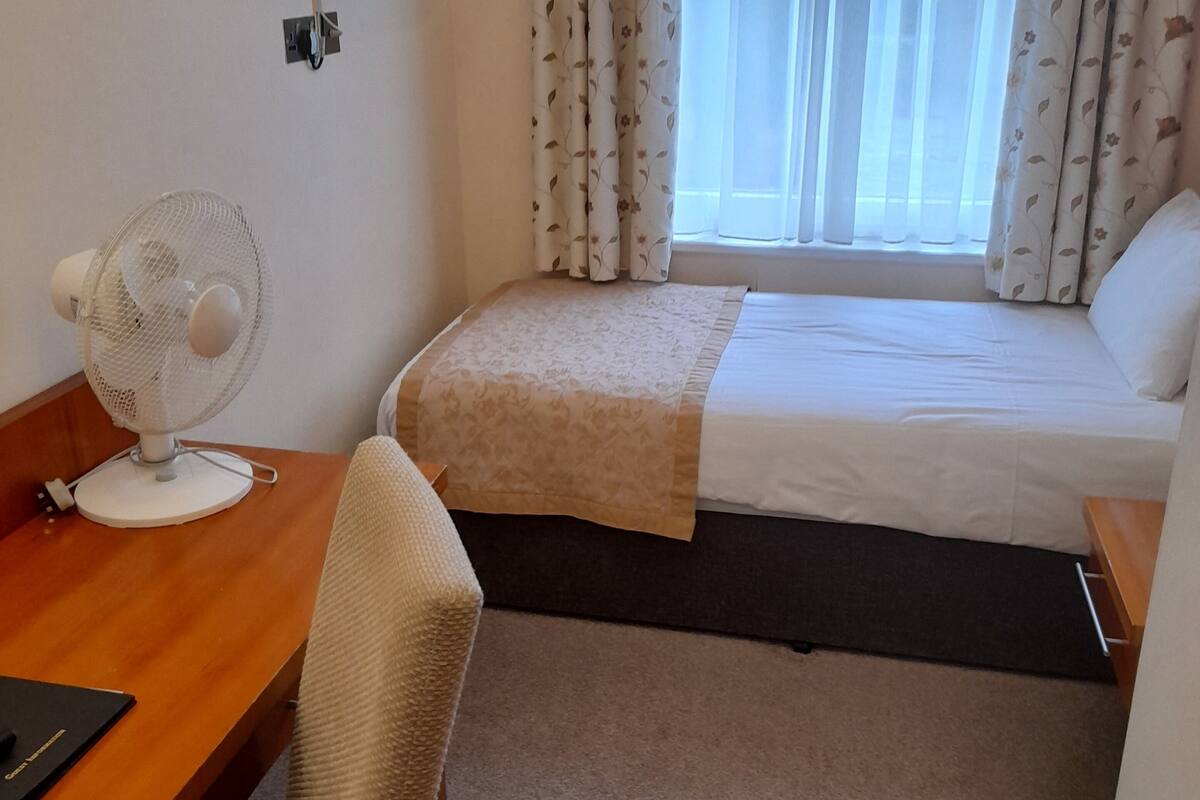 A hotel room with a bed, a desk with a chair, a standing fan, a TV mounted on the wall, and a window with curtains.