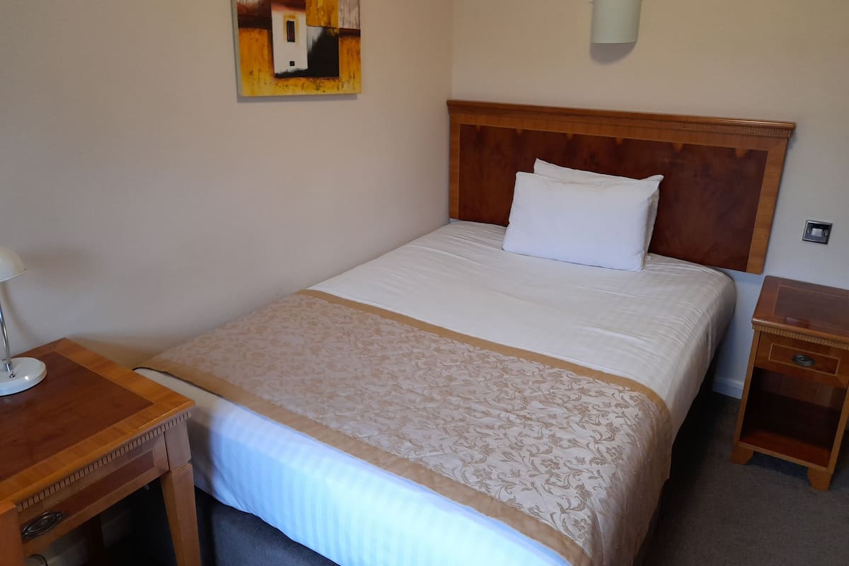 A neatly made bed with a wooden headboard, a bedside table with a lamp, and a small wooden nightstand.