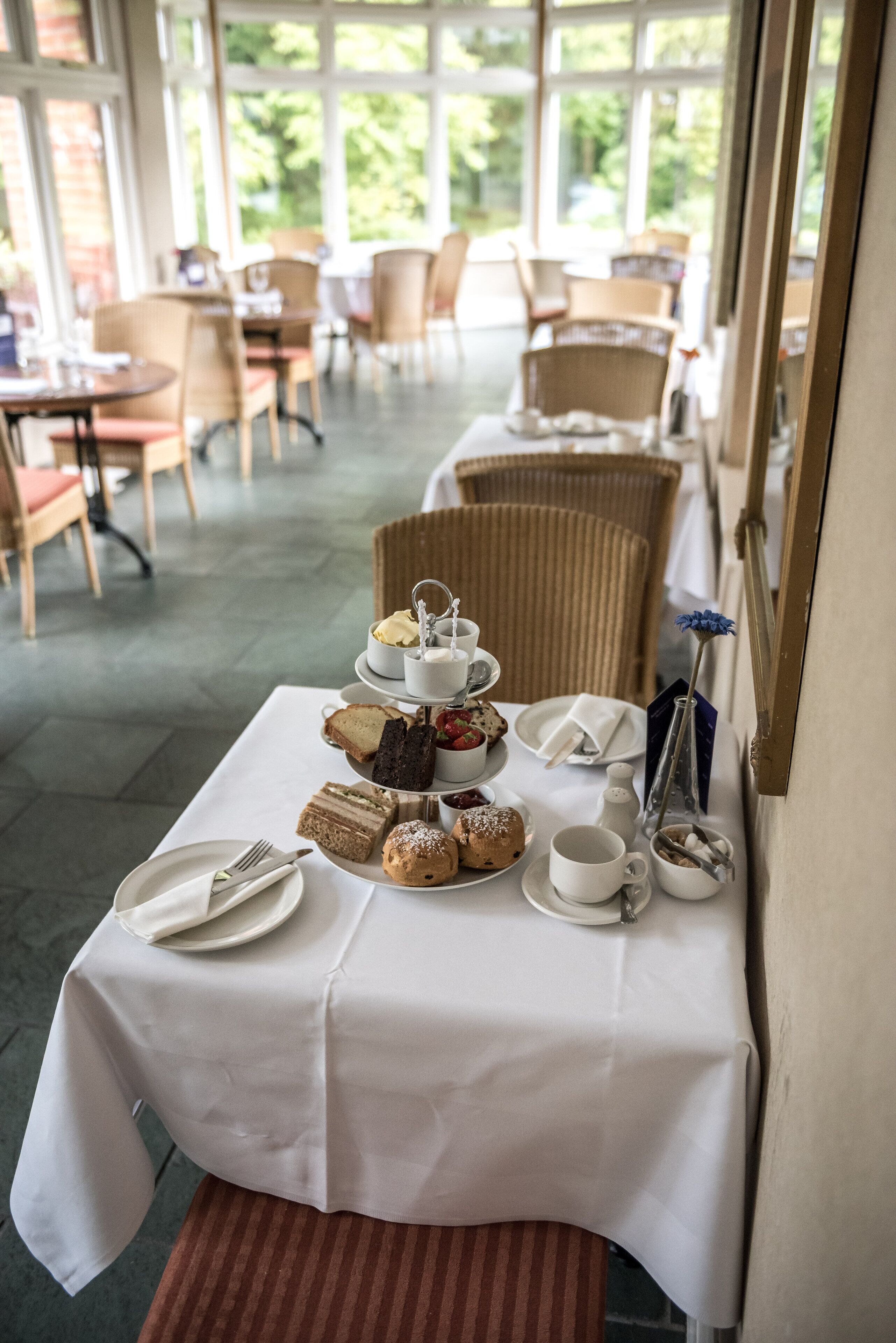 Un tavolo apparecchiato con una varietà di pasticcini e dolci, disposti su una tovaglia bianca in una sala da pranzo luminosa con sedie in vimini e ampie finestre.