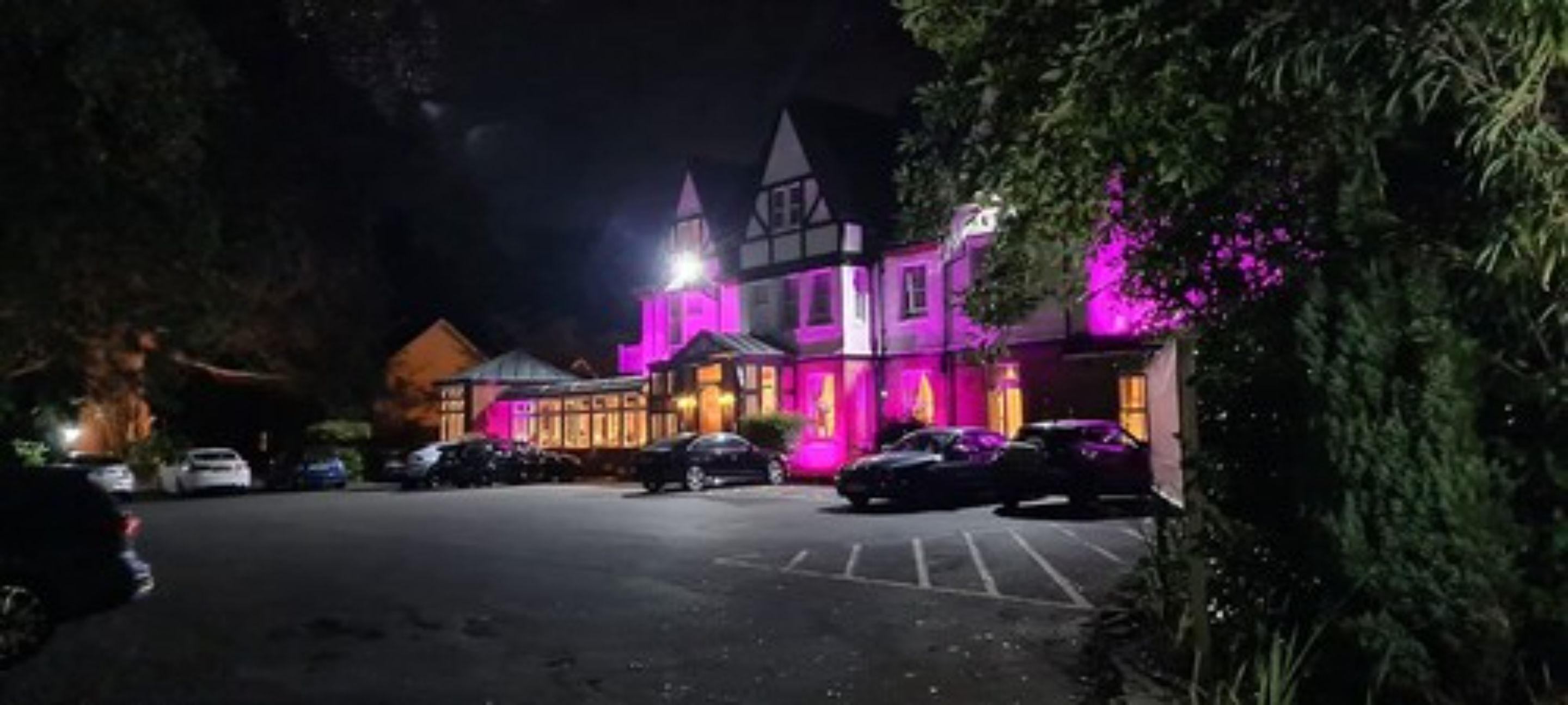 A building with a Tudor-style architecture, illuminated with purple lights, featuring a parking lot and a few parked cars.