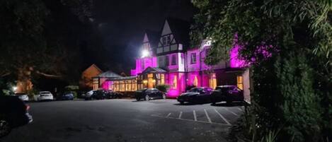 A building with a Tudor-style architecture, illuminated with purple lights, featuring a parking lot and a few parked cars.
