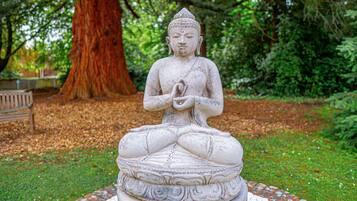 Una estatua de Buda de piedra en un jardín con un árbol grande y un banco al fondo.