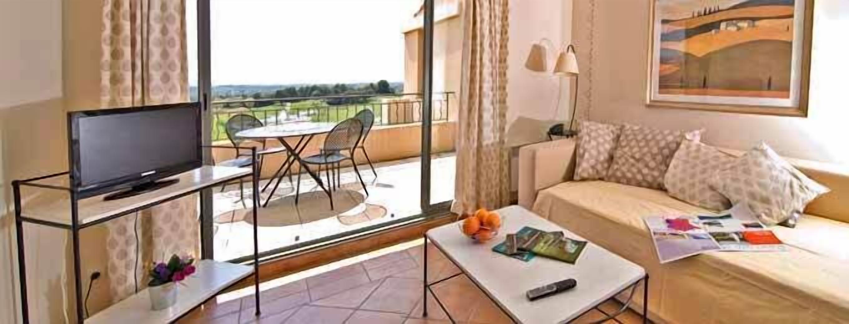 A modern hotel room with a balcony, a sofa, a coffee table, and a TV on a glass stand.