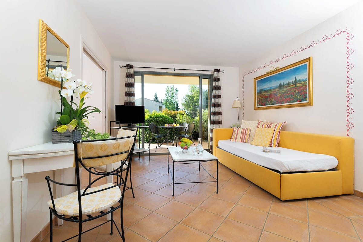 A living room with a yellow sofa, a TV, a dining table with chairs, a plant, and a painting on the wall.