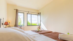 A hotel room with two beds, a window with curtains, a lamp, and a view of greenery outside.