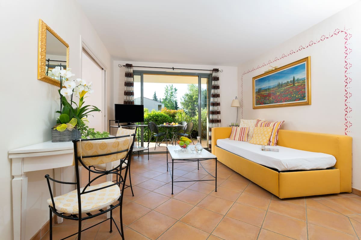 A living room with a yellow sofa, a dining table with chairs, a TV, and a window with curtains.