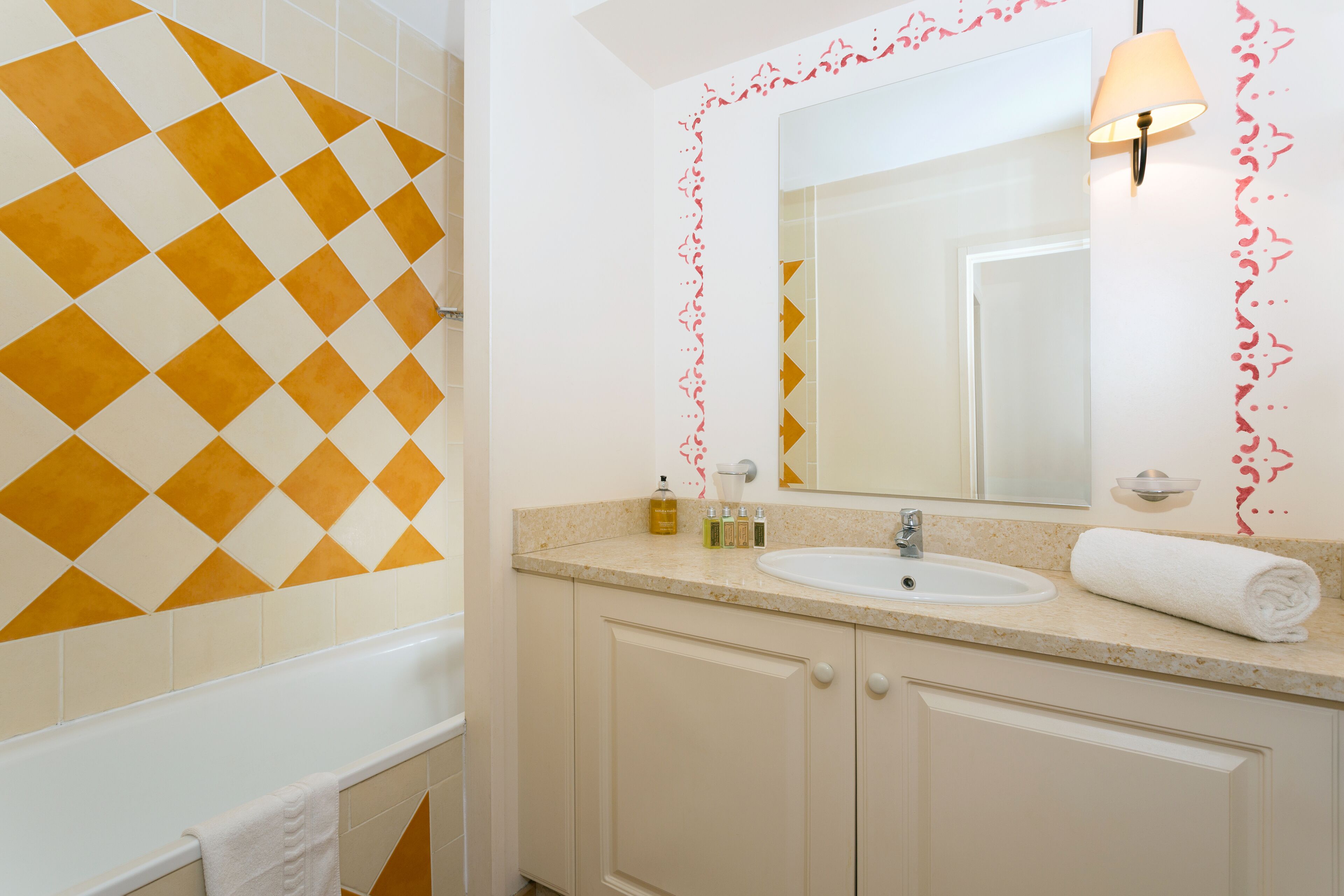 A bathroom with a white sink, mirror, and beige cabinets. There is a towel on the edge of the tub and toiletries on the counter.