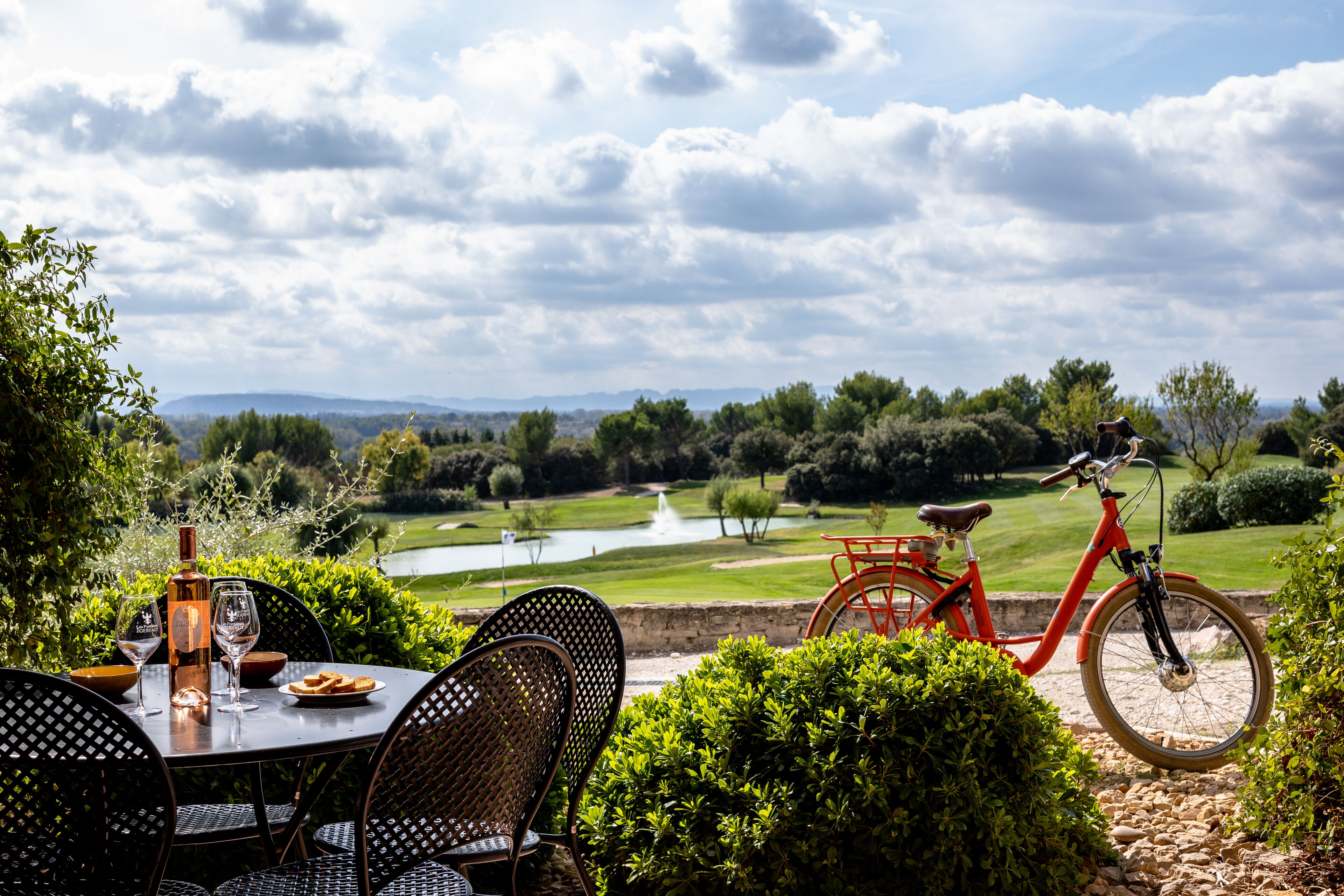 Mesa con sillas, una botella y vasos, con vista a un campo de golf y una bicicleta roja.