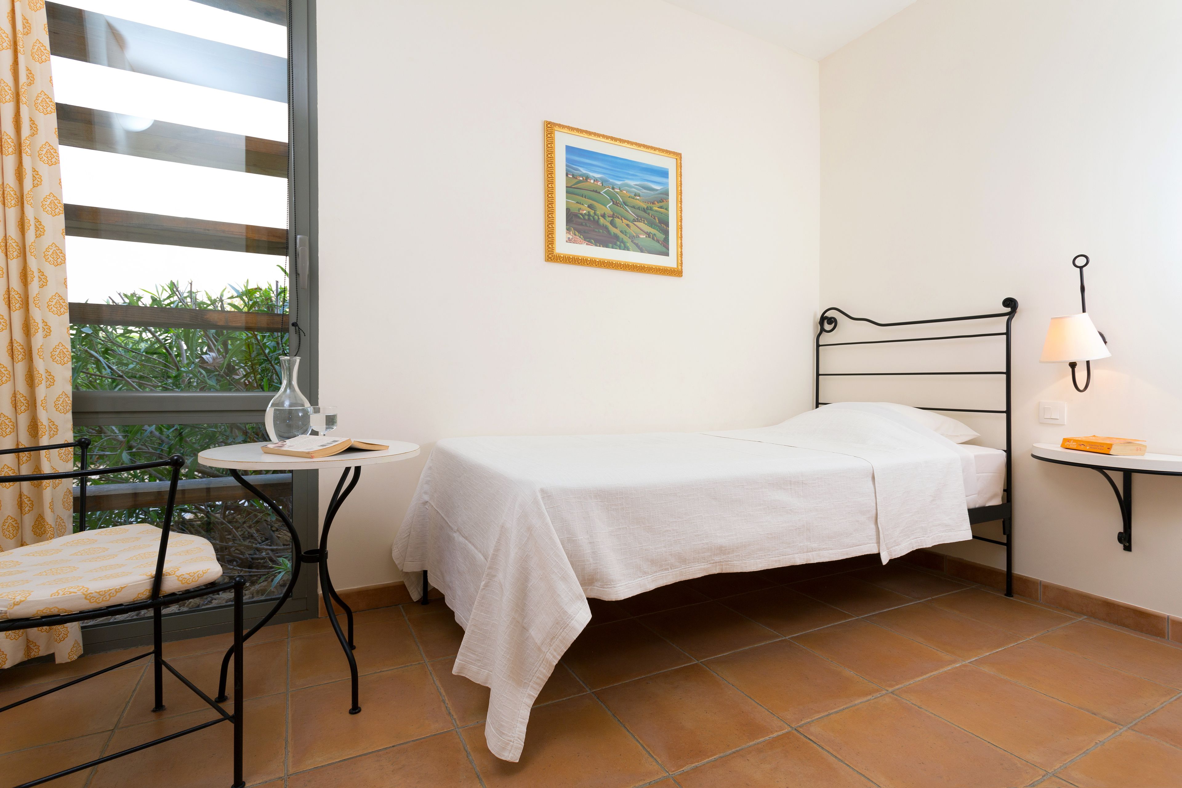 Una habitación pequeña y ordenada con una cama, una mesa y una silla. Cuenta con un ventanal con cortinas, un cuadro enmarcado en la pared y una lámpara sobre una mesita.