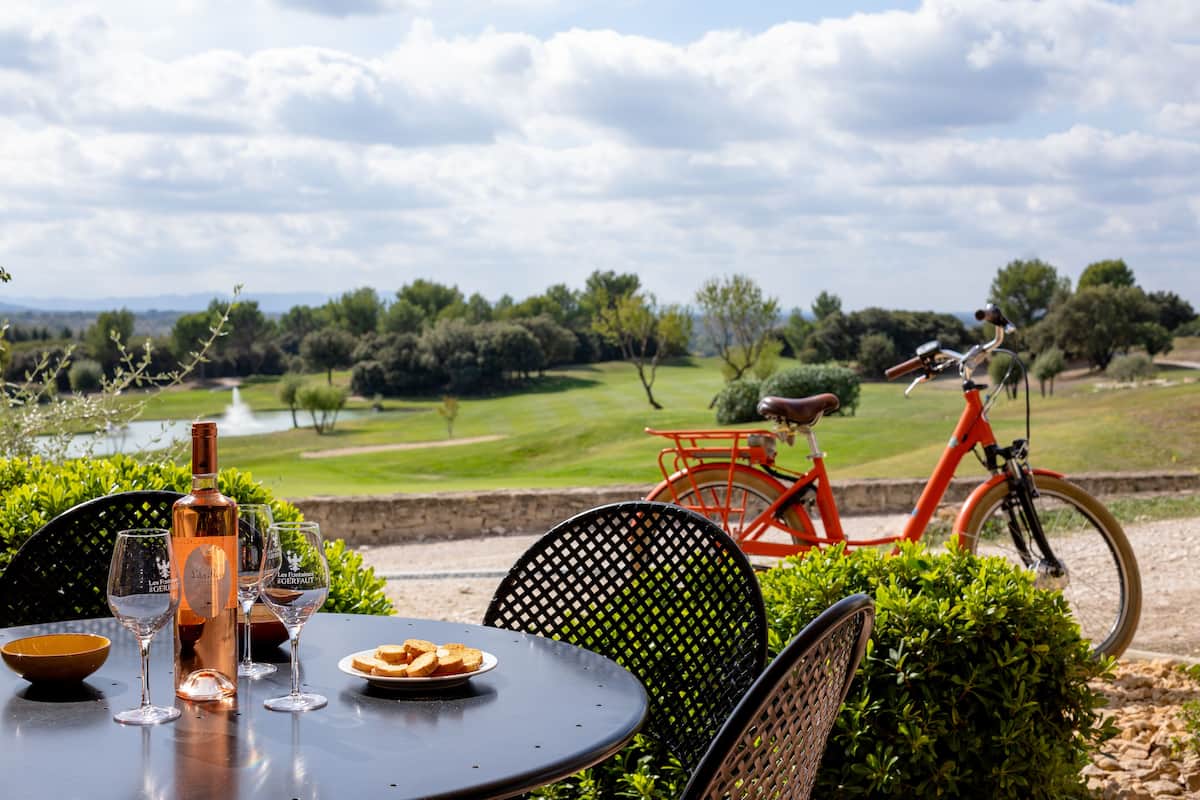 A table with wine glasses, a bottle, and a plate of food, set outdoors with a bicycle and greenery in the background.