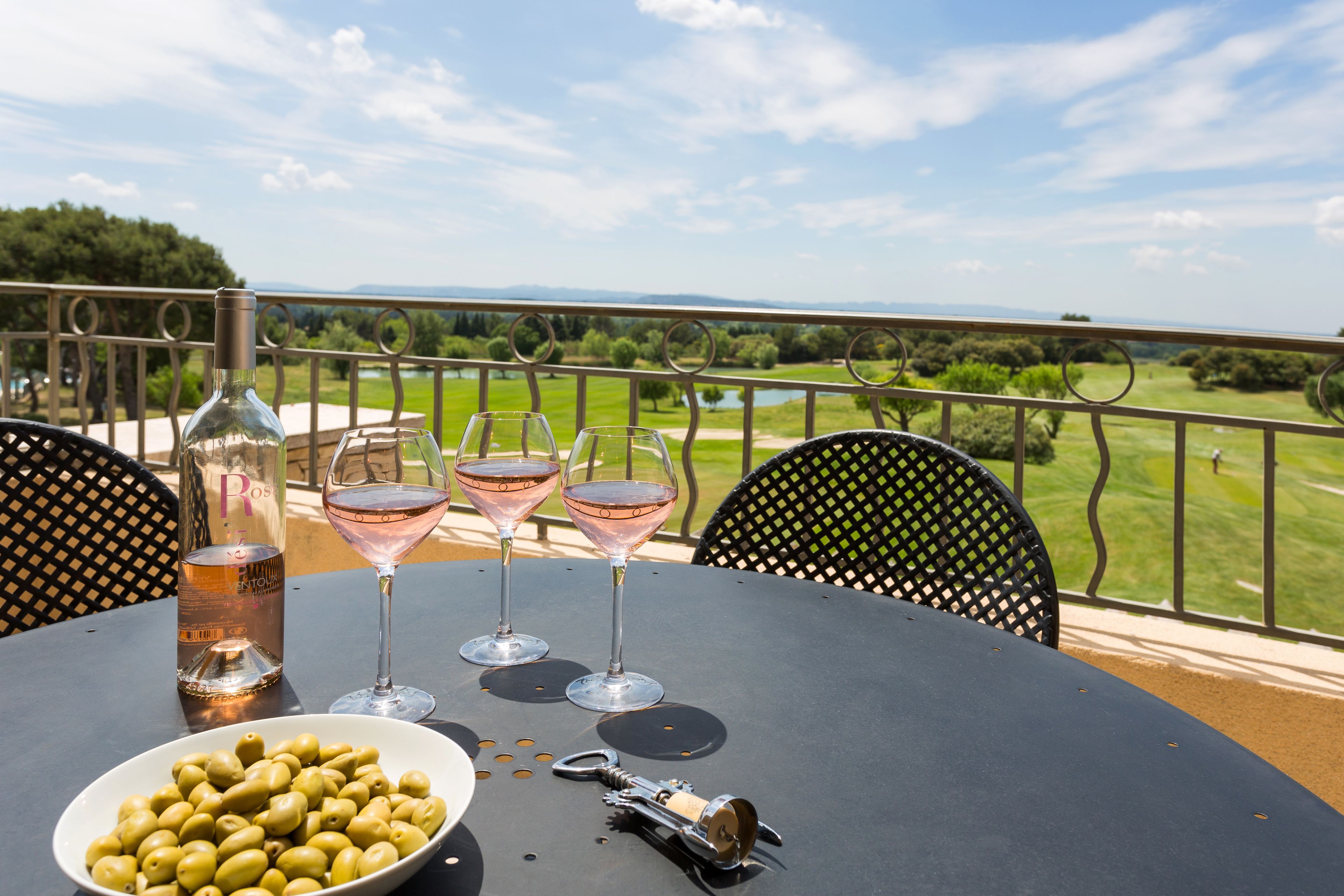 A table with a bottle of rosé wine, two glasses, a bowl of olives, and a corkscrew on a balcony overlooking a golf course.