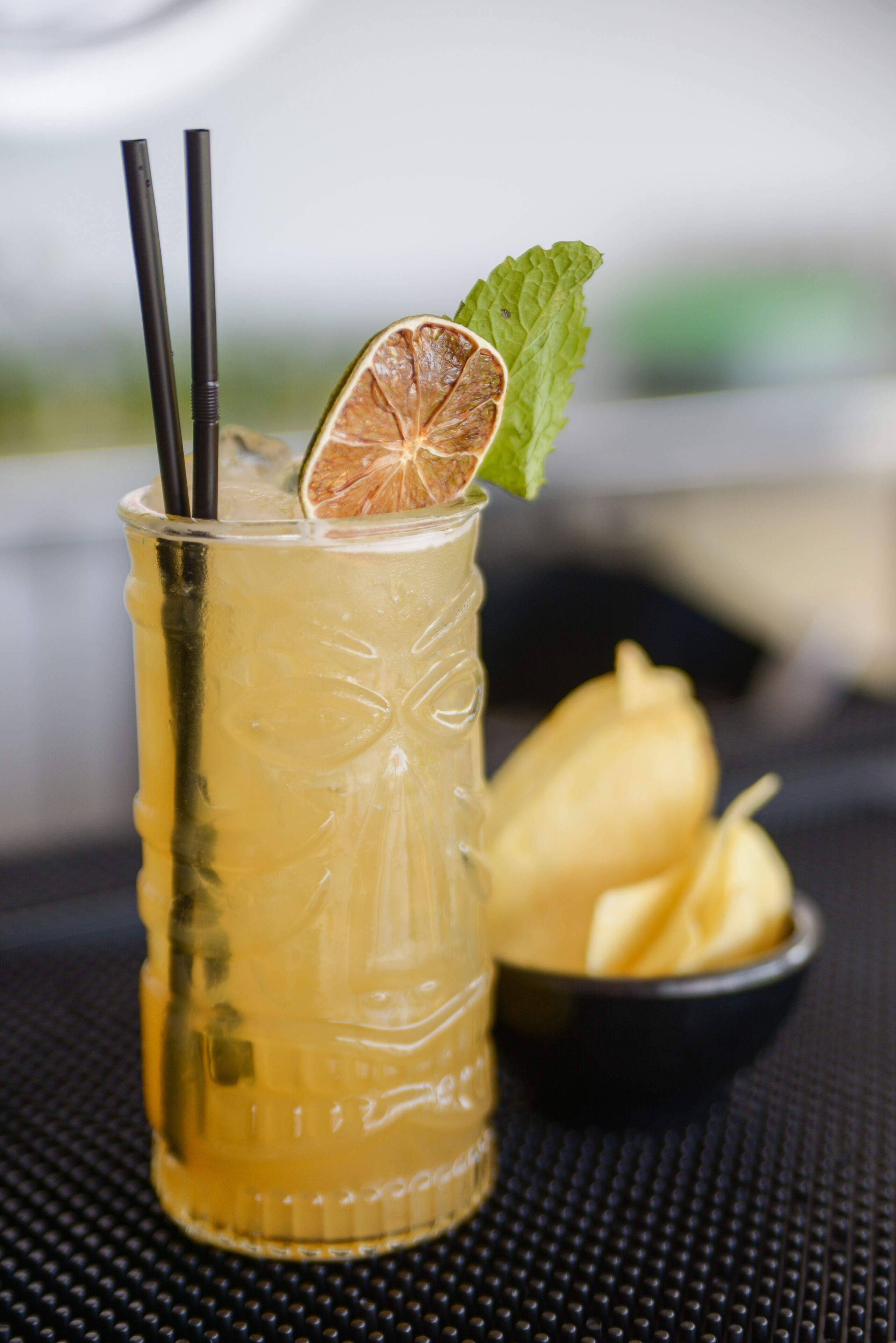 A glass of yellow beverage with a lime slice and mint leaf, accompanied by a bowl of yellow fruit.