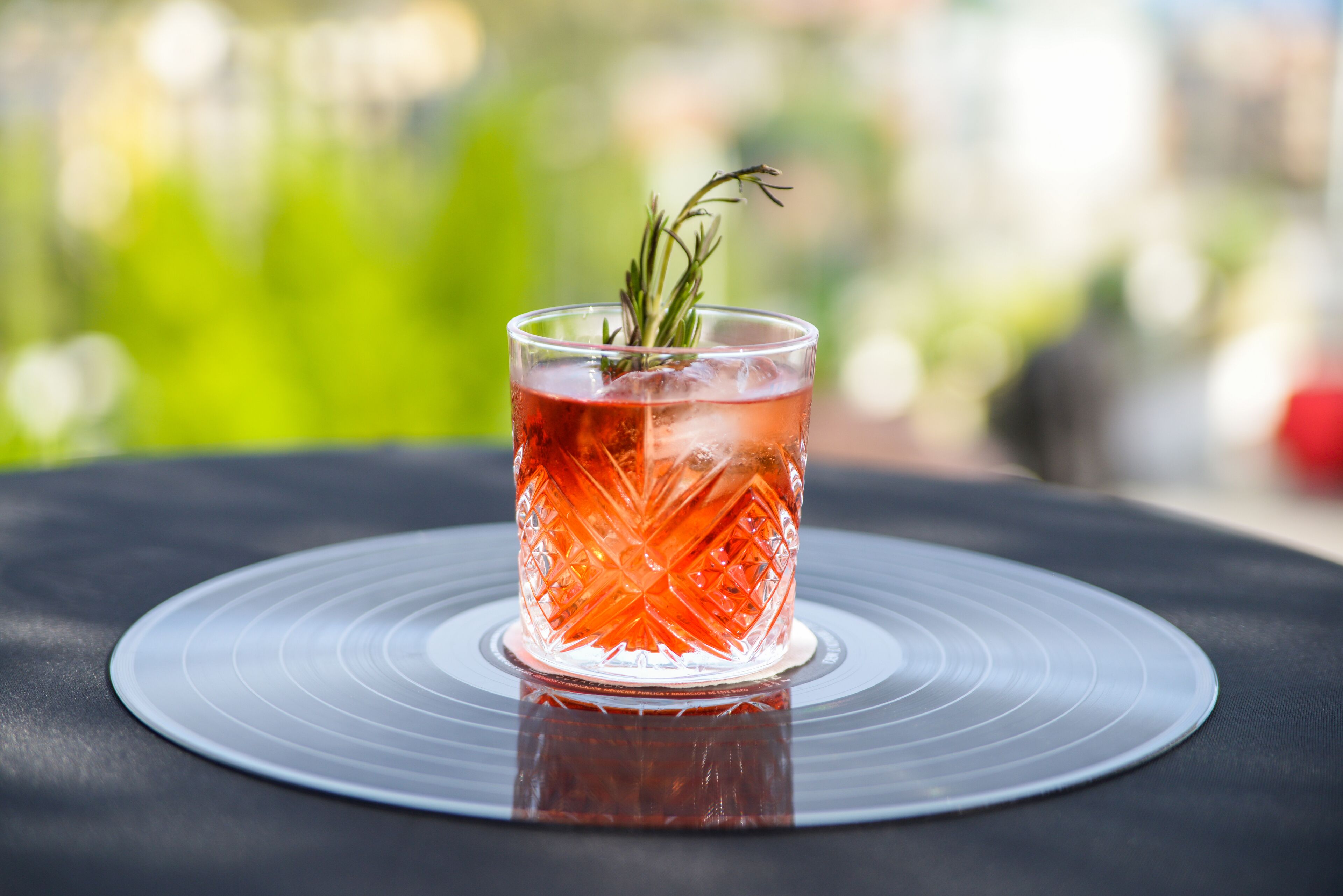 Un verre transparent contenant une boisson rouge, agrémenté d’une branche de romarin, posé sur une table noire ornée d’un motif circulaire.