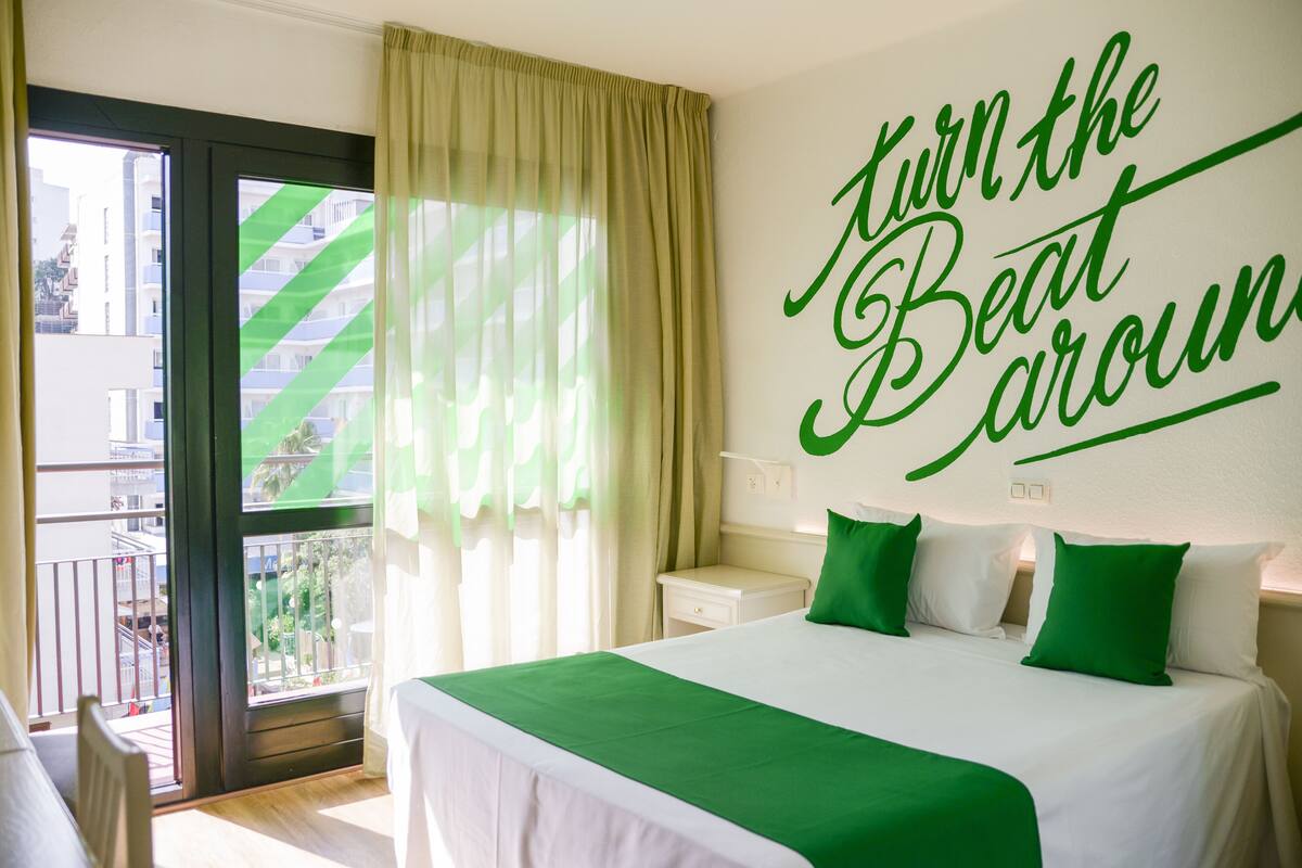 A hotel room with a bed, a nightstand, a window with curtains, and a wall with a green sign.