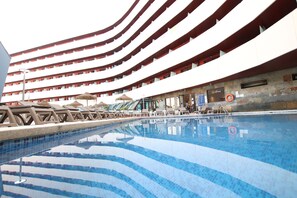 Une piscine entourée de chaises longues et de parasols, elle-même bordée d’un immeuble à plusieurs étages doté de balcons.