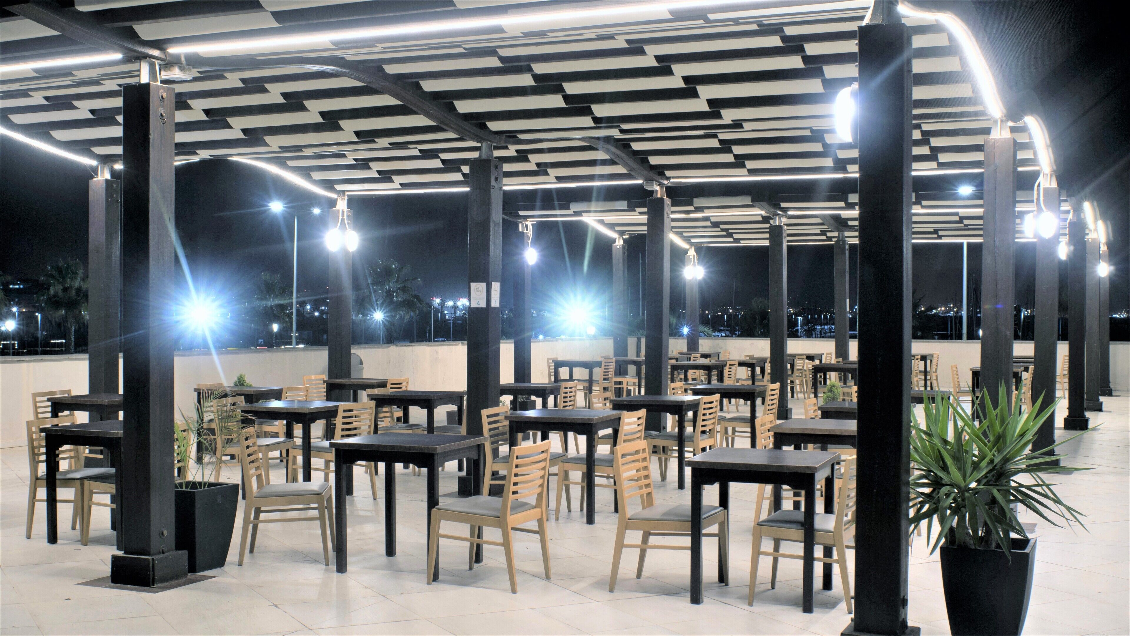 Un área de comedor al aire libre con mesas y sillas, piso de baldosas y techo con luminarias lineales.