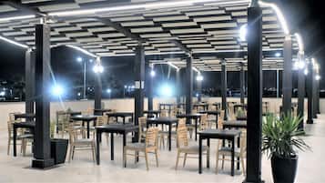 Un área de comedor al aire libre con mesas y sillas, piso de baldosas y techo con luminarias lineales.