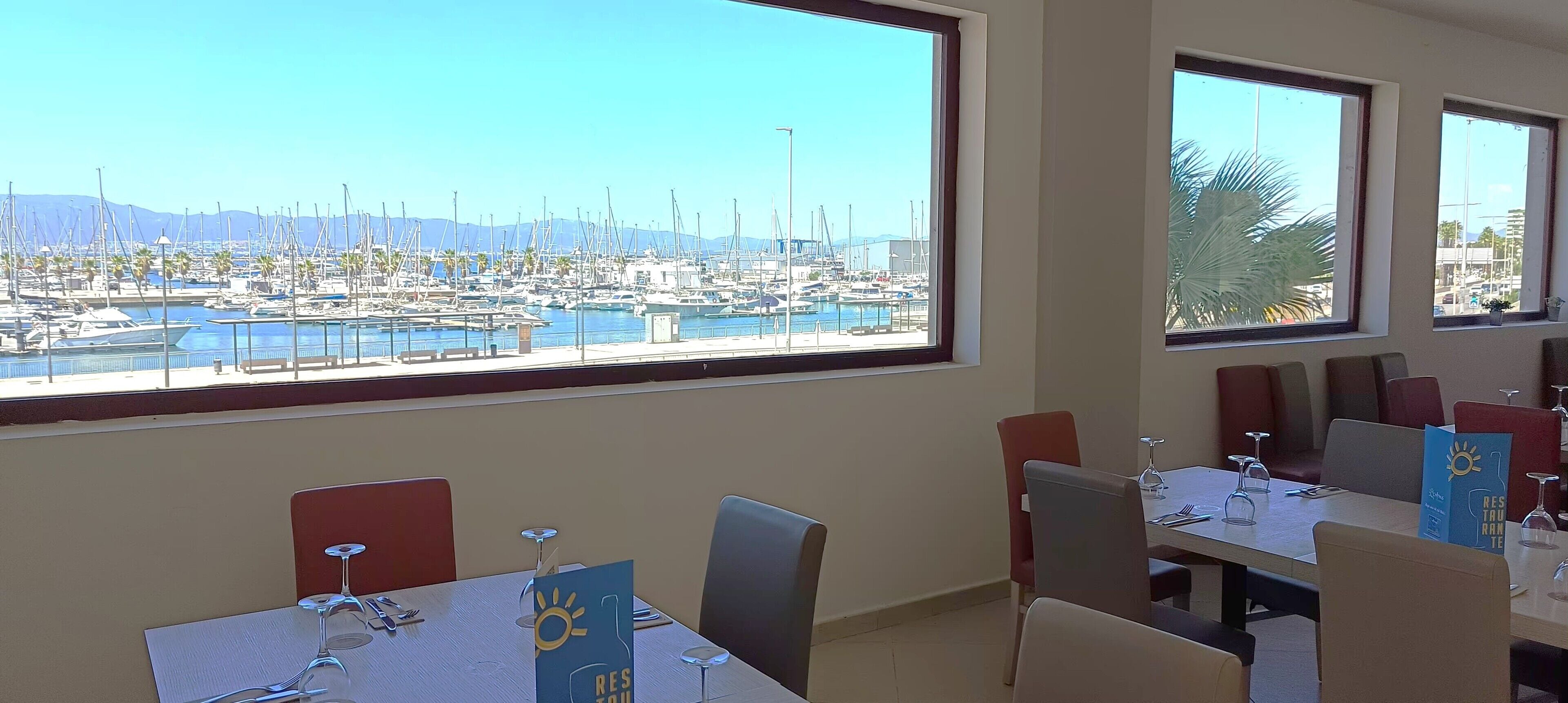 Un espace repas avec des tables préparées pour deux personnes, de grandes fenêtres donnant sur le port de plaisance et un panneau indiquant la présence d’une terrasse de restaurant.