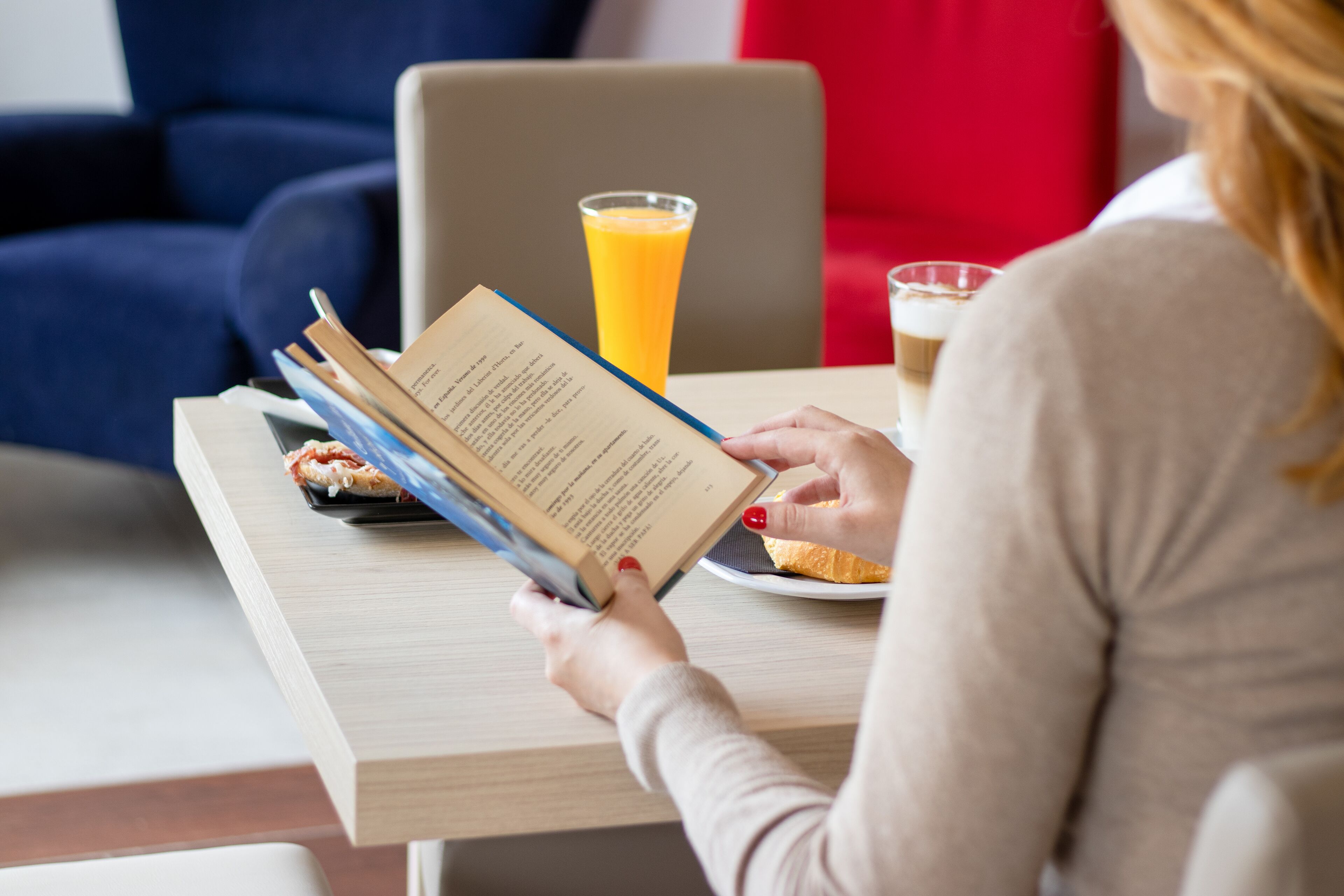 Una persona leyendo un libro en una mesa con un vaso de jugo de naranja y un plato de comida.