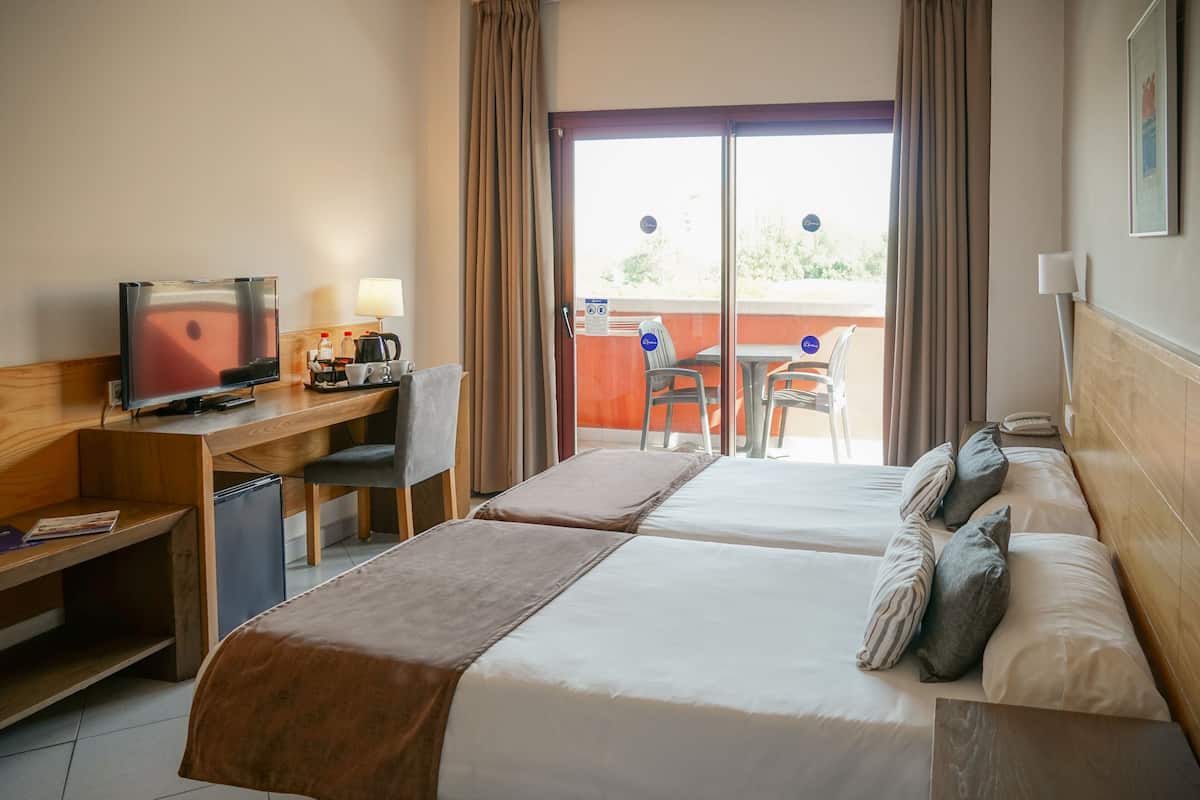 A hotel room with two beds, a desk with a television, a coffee maker, and a view of the outside balcony.