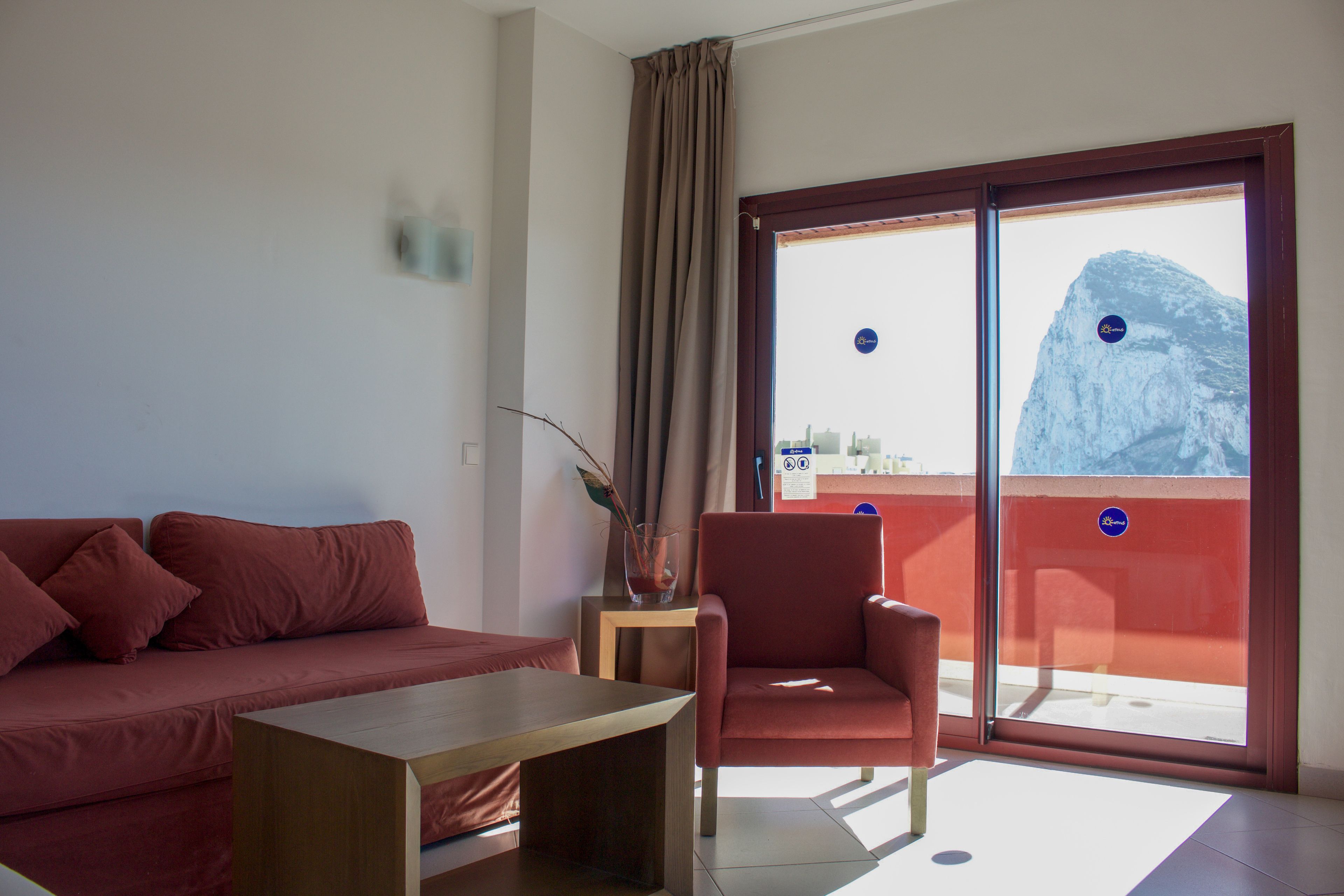 Ein Zimmer mit einem roten Sofa, einem Holztisch und einem roten Sessel. Ein Fenster bietet Blick auf einen Berg.