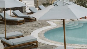A poolside area with lounge chairs and umbrellas, stone flooring, and a pool. - Argo Hotel (Mykonos)