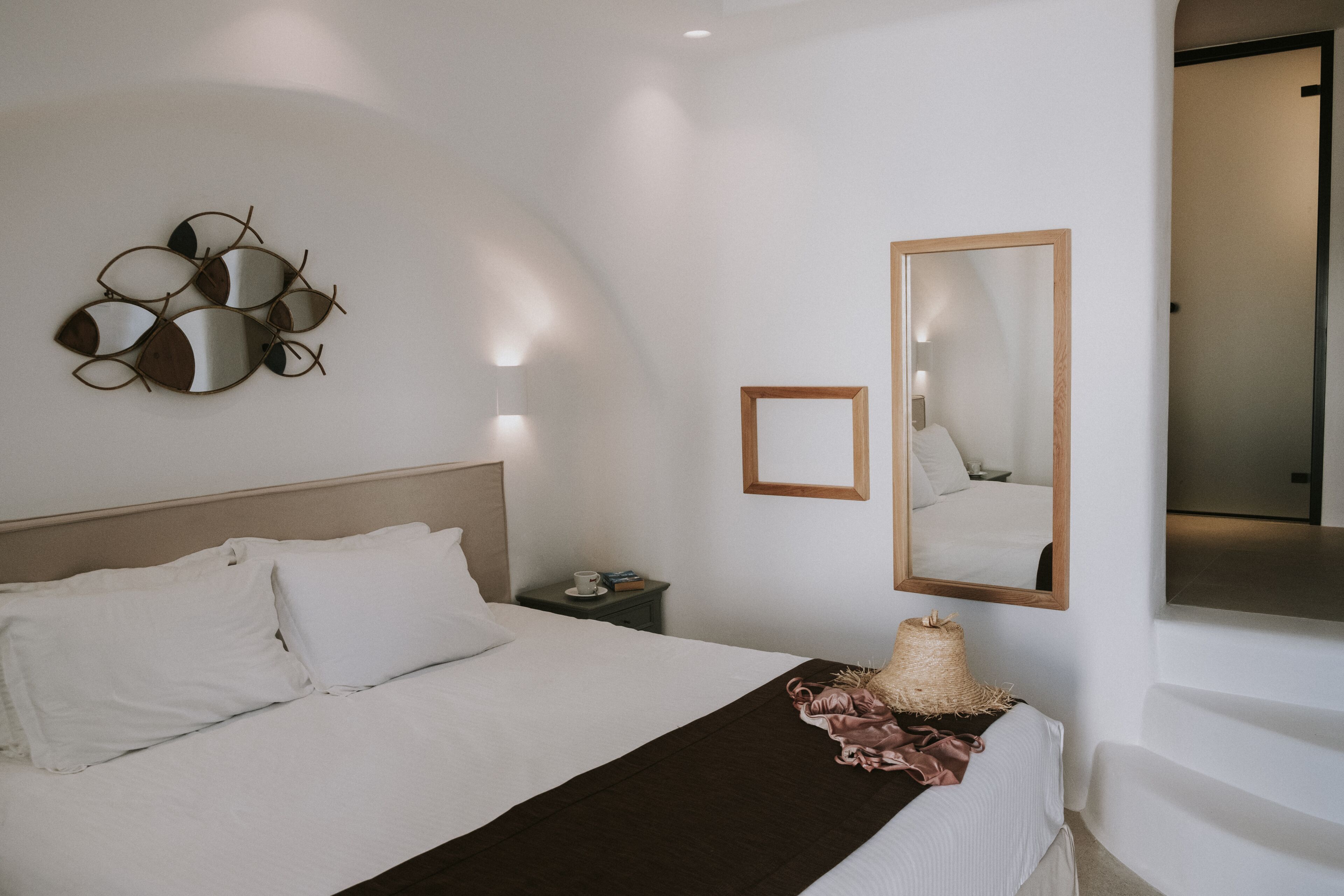 A neatly made bed with white linens, a bedside table with a cup and a book, a wall-mounted mirror, and a decorative wall art piece.