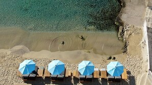 An aerial view of a beach with clear turquoise water, a sandy shore, and wooden lounge chairs with blue umbrellas arranged in a row. - Argo Hotel (Mykonos)