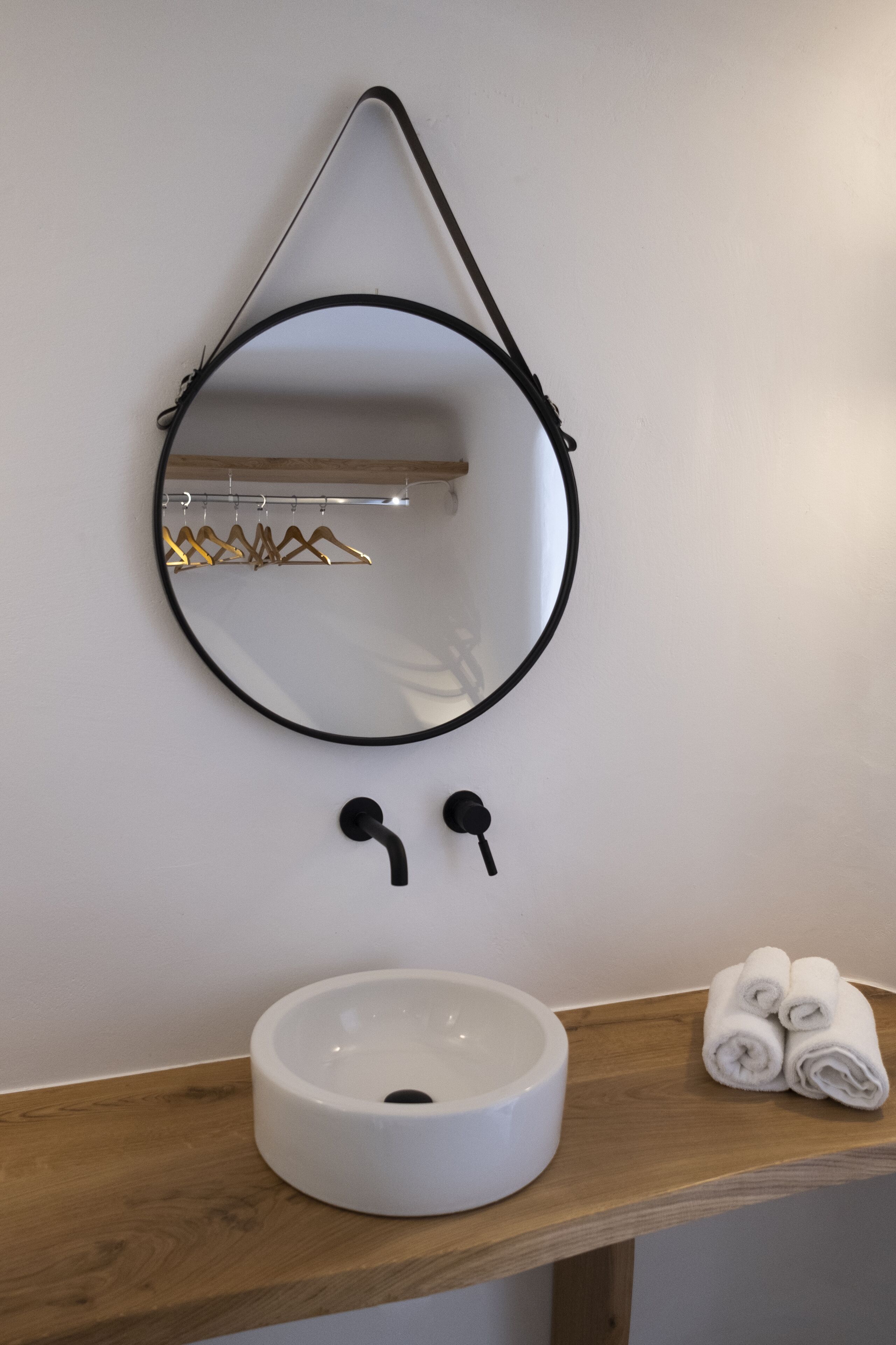 Une salle de bain avec un miroir rond, un lavabo blanc et une étagère en bois où sont posées des serviettes.