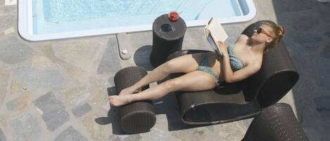 A person relaxing by a swimming pool with a book, surrounded by stone flooring and wicker chairs.
