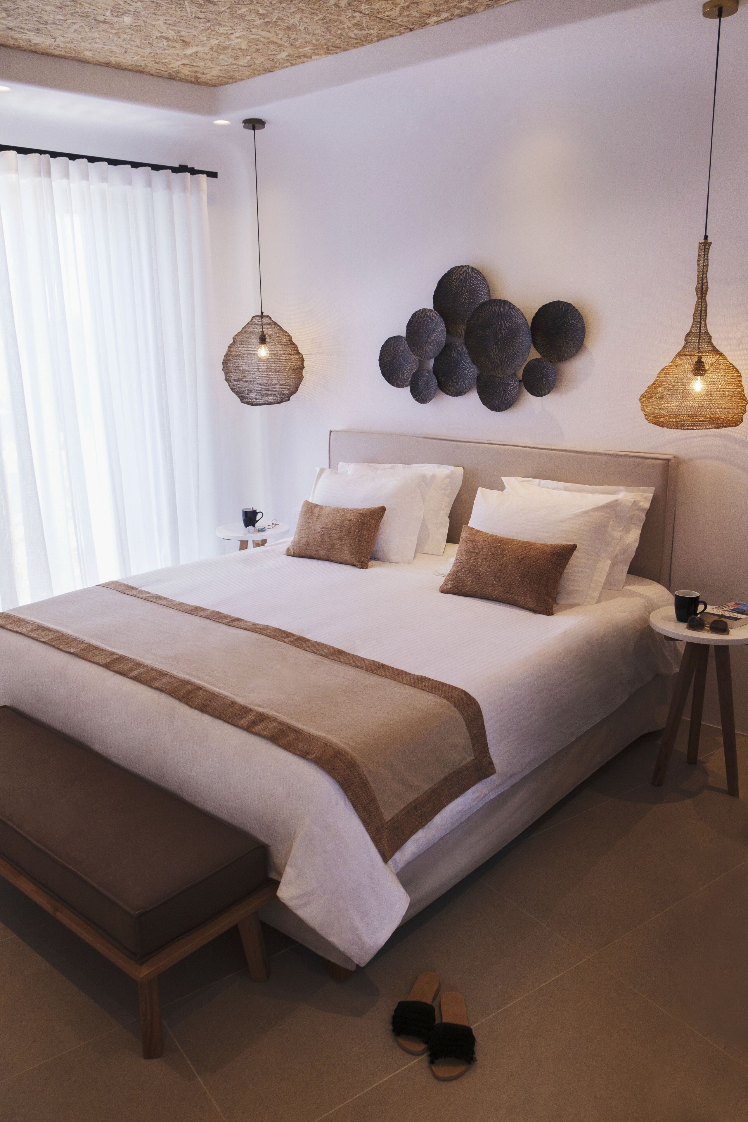 A neatly made bed with a brown bench, a small bedside table with a cup and a book, and a wall-mounted light fixture.