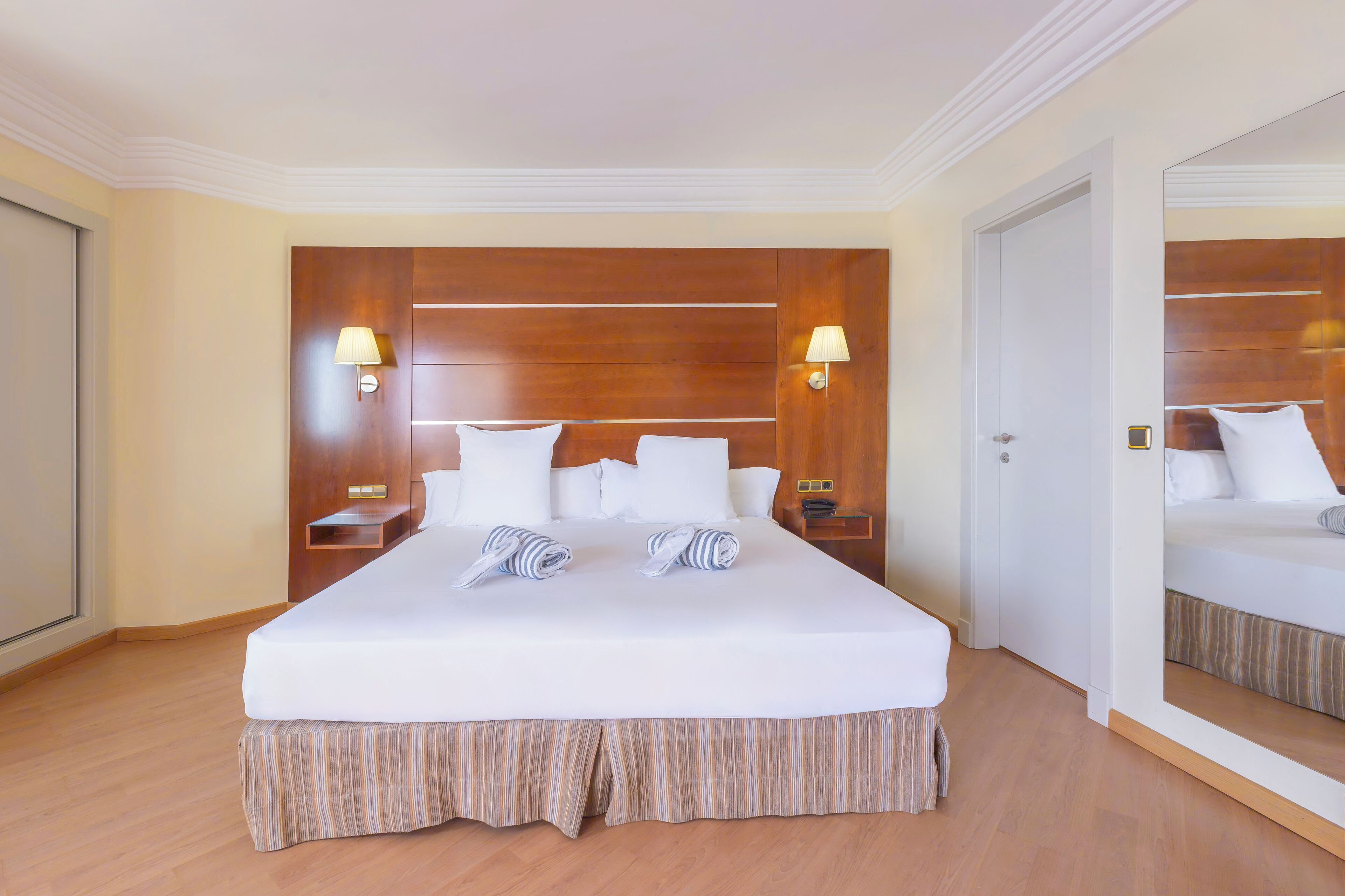 A hotel room with a large bed, wooden headboard, two bedside tables with lamps, and a mirror on the wall.