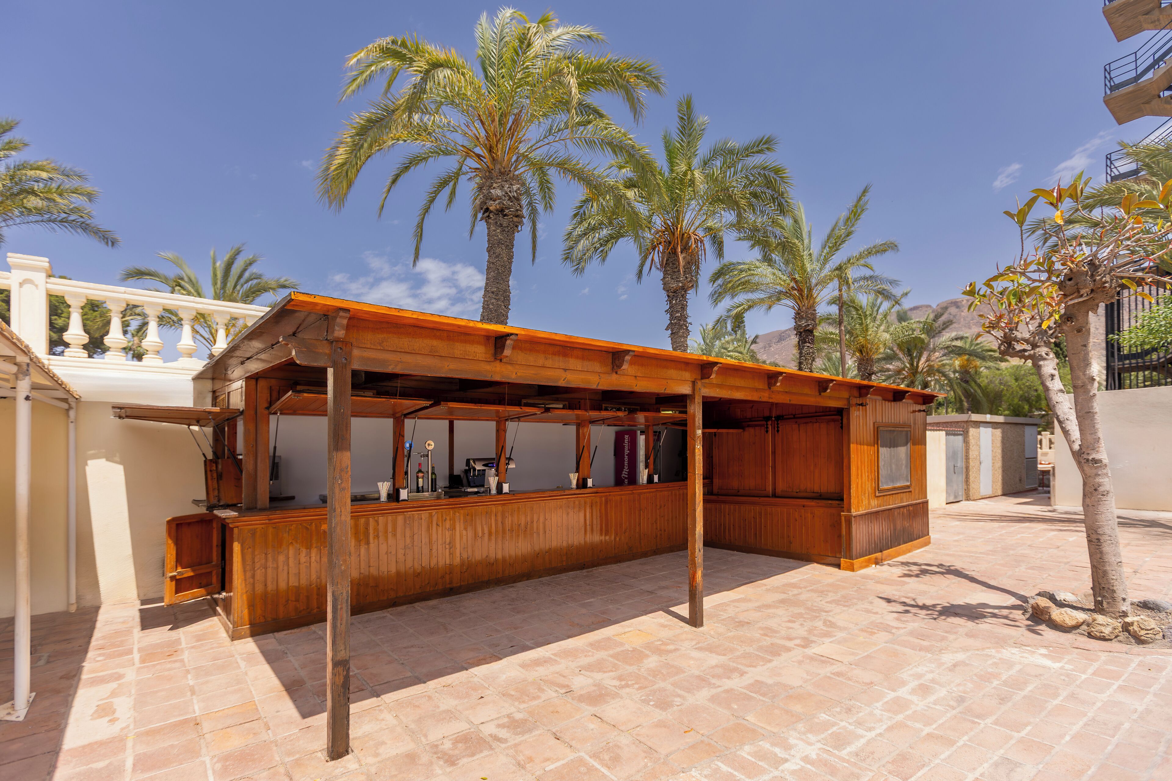 Un área de bar al aire libre de madera con piso de baldosas, rodeada de palmeras y un edificio blanco con balcón.