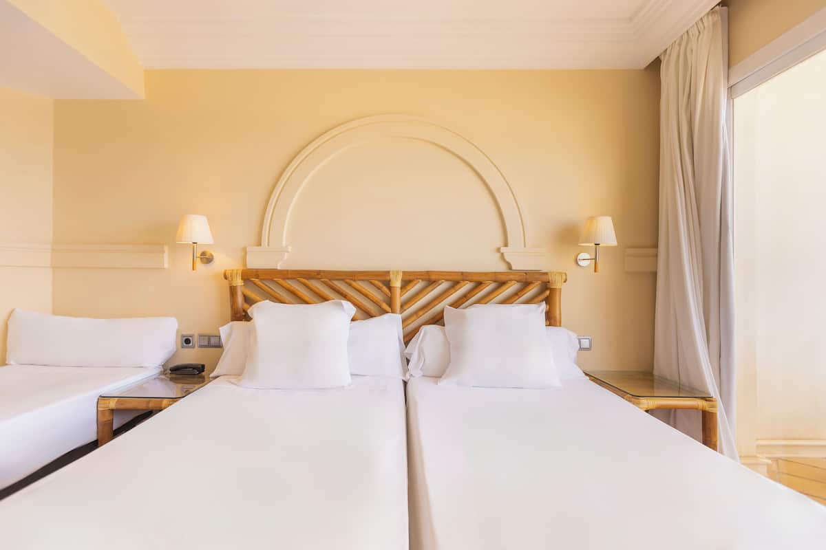 Una habitación de hotel con dos camas individuales, un cabecero de madera, lámparas de noche y un gran ventanal con cortinas.