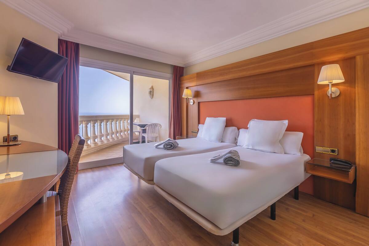 Habitación de hotel con dos camas, televisión, balcón y cabecero de madera.