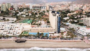 Un paisaje urbano costero con un hotel destacado de varios pisos, una playa en primer plano y montañas al fondo.
