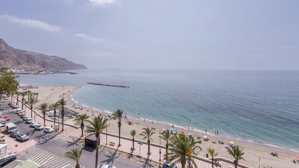 Una carretera costera con coches aparcados, palmeras y una playa con gente y sombrillas.
